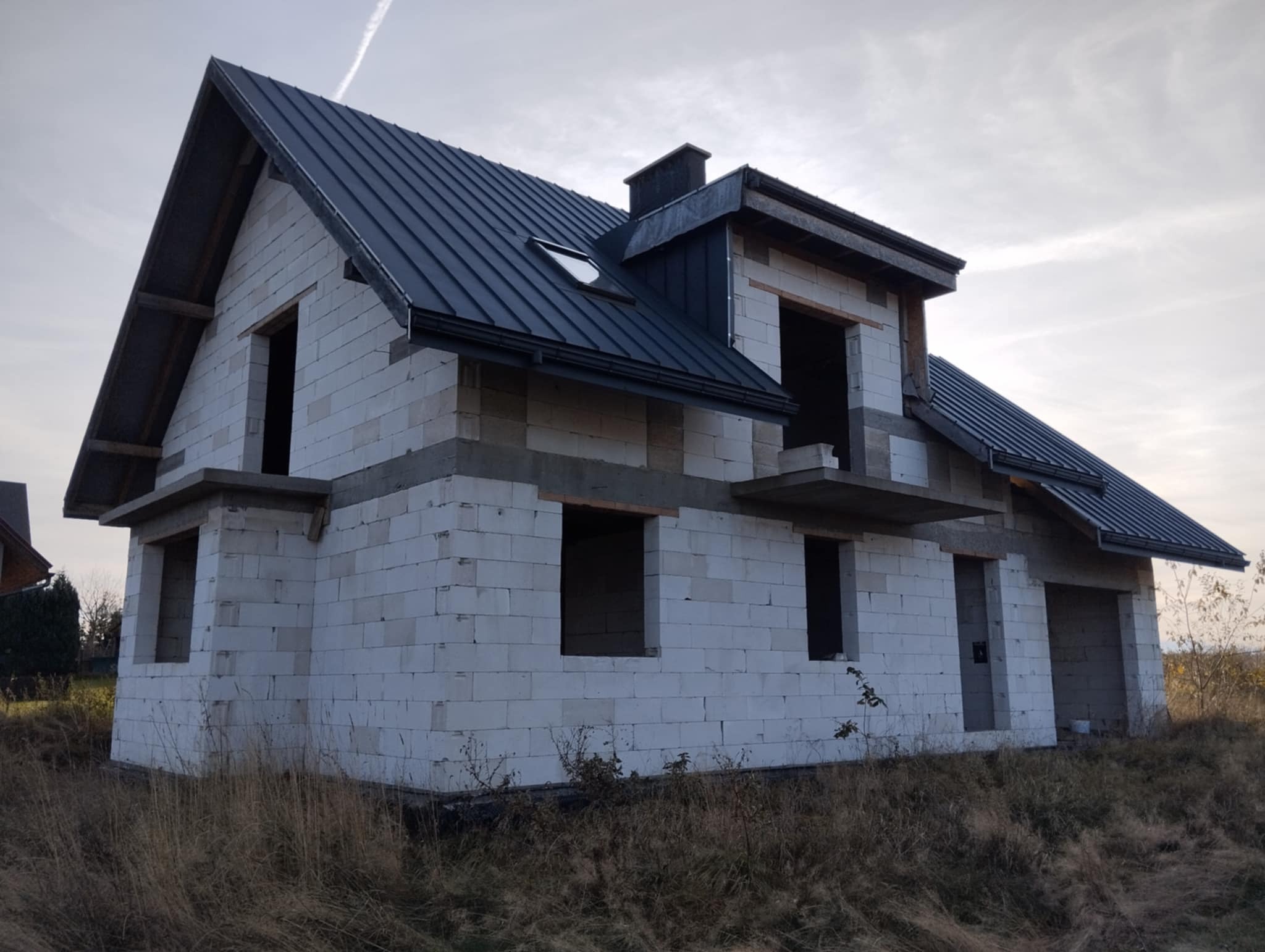 Surowy stan domu z białych pustaków z zamontowanym, grafitowym dachem z blachy trapezowej i rynnami, widoczne okna dachowe i komin, otoczenie działki z suchą trawą.