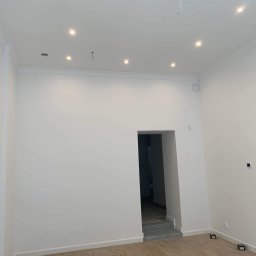 Kamil Wolter - Pomalowane na biało wnętrze z drewnianą podłogą, oświetleniem punktowym w suficie i otwartym przejściem do innego pomieszczenia z szarymi schodami i podłogą.