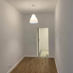 Pomalowane na biało wnętrze z drewnopodobnymi panelami podłogowymi, widoczny fragment korytarza z szaro-białą wzorzystą podłogą, biała lampa wisząca i regulator temperatury na ścianie.