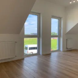 Jasne panele podłogowe ułożone w pustym pokoju na poddaszu z dwoma oknami, przez które widać zielone pole i biały bus.