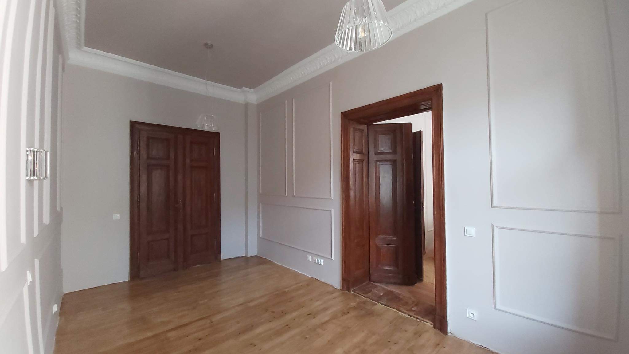 Wnętrze pokoju we Wrocławiu po odświeżeniu, z drewnianą podłogą, dwojgiem ciemnych drewnianych drzwi, ścianami w kolorze złamanej bieli z dekoracyjnymi panelami i listwami przysufitowymi...