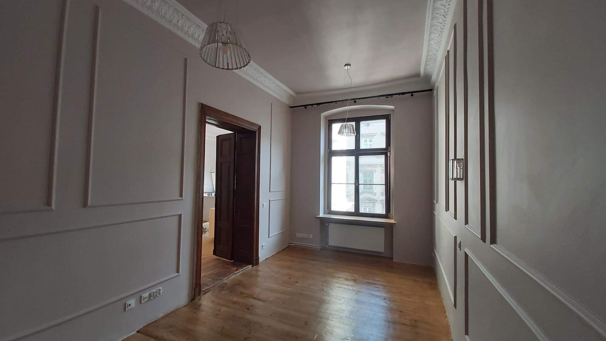 Pokój po remoncie z drewnianą podłogą, szarymi ścianami z dekoracyjnymi listwami, oknem z ciemną ramą i otwartymi drewnianymi drzwiami do sąsiedniego pomieszczenia.