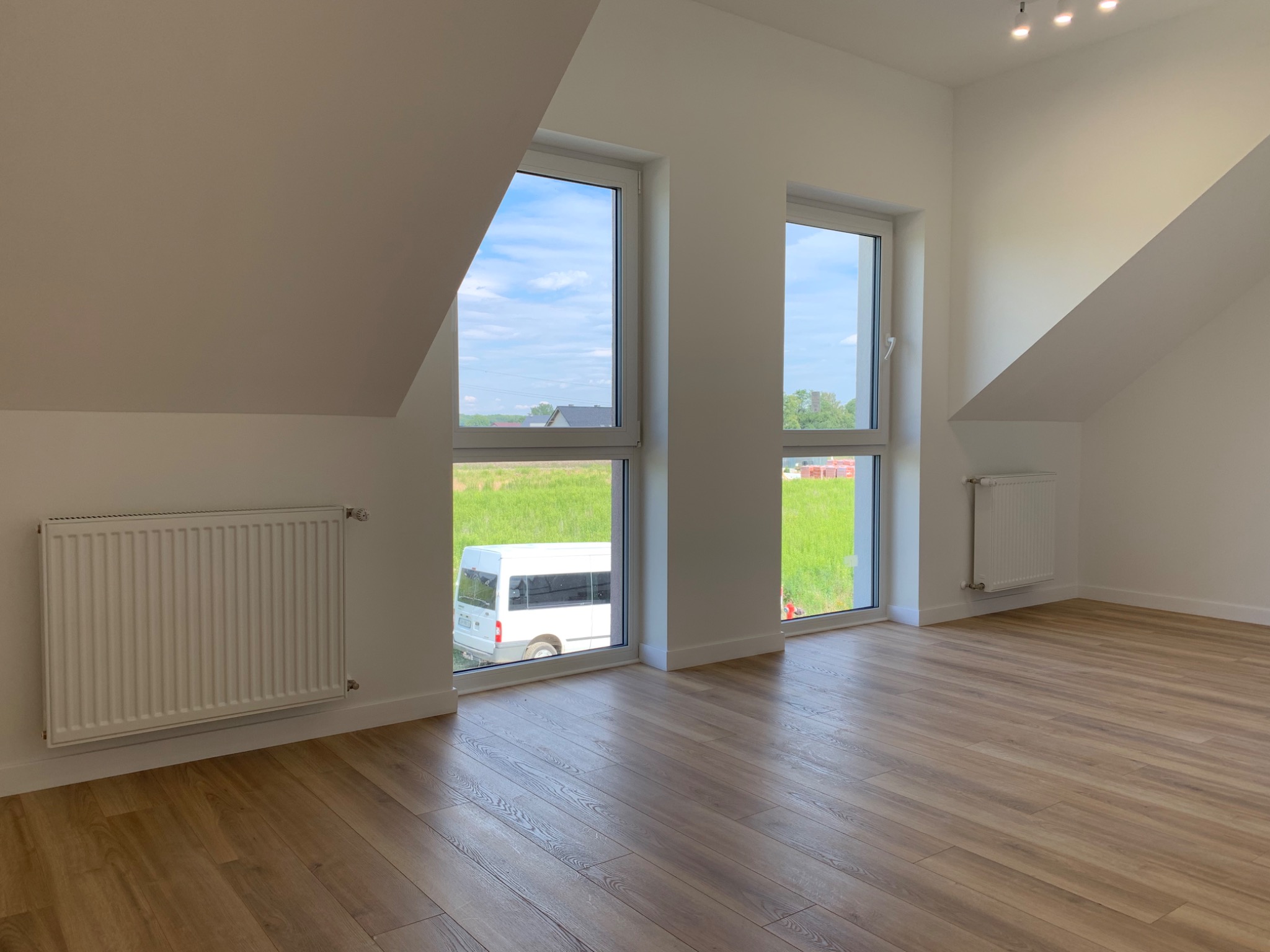 Jasne panele podłogowe ułożone w pustym pokoju na poddaszu z dwoma oknami, przez które widać zielone pole i biały bus.