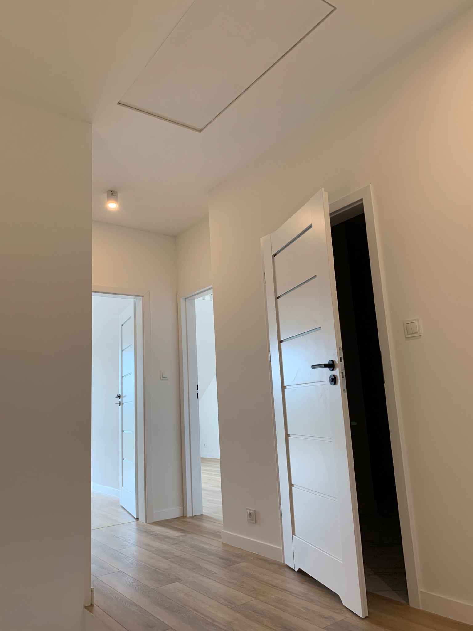 Jasne wnętrze korytarza z panelami podłogowymi w odcieniach drewna, białymi ścianami i drzwiami z poziomymi frezami, otwartymi do wnętrza, oraz klapą na strych w suficie.