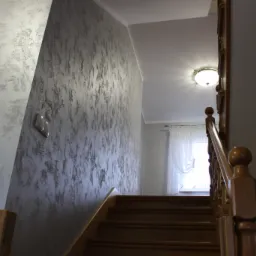 Ściana w klatce schodowej z dekoracyjną, strukturalną gładzią w odcieniach szarości, widoczne schody drewniane i balustrada, w tle okno z firanką.