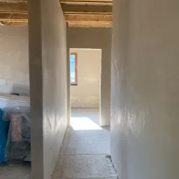 Korytarz w trakcie prac wykończeniowych, świeżo nałożone tynki na ścianach, surowy jastrych na podłodze, widoczne drewniane elementy konstrukcji stropu, okno w oddali.