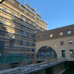 Budynek w trakcie remontu elewacji, z rusztowaniami i zabezpieczonymi oknami, widoczny fragment kanału wodnego.