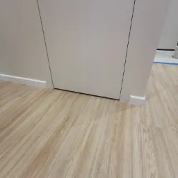 Jasne panele podłogowe ułożone wzdłuż białej ściany z drzwiami. Widoczne listwy przypodłogowe. Minimalistyczny design wnętrza.