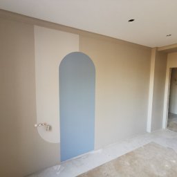 Adamas-Wr - Wnętrze po odświeżeniu we Wrocławiu. Ściana z geometrycznym, niebieskim wzorem łuku, jasne kolory, widoczne gniazdka elektryczne i surowa podłoga.