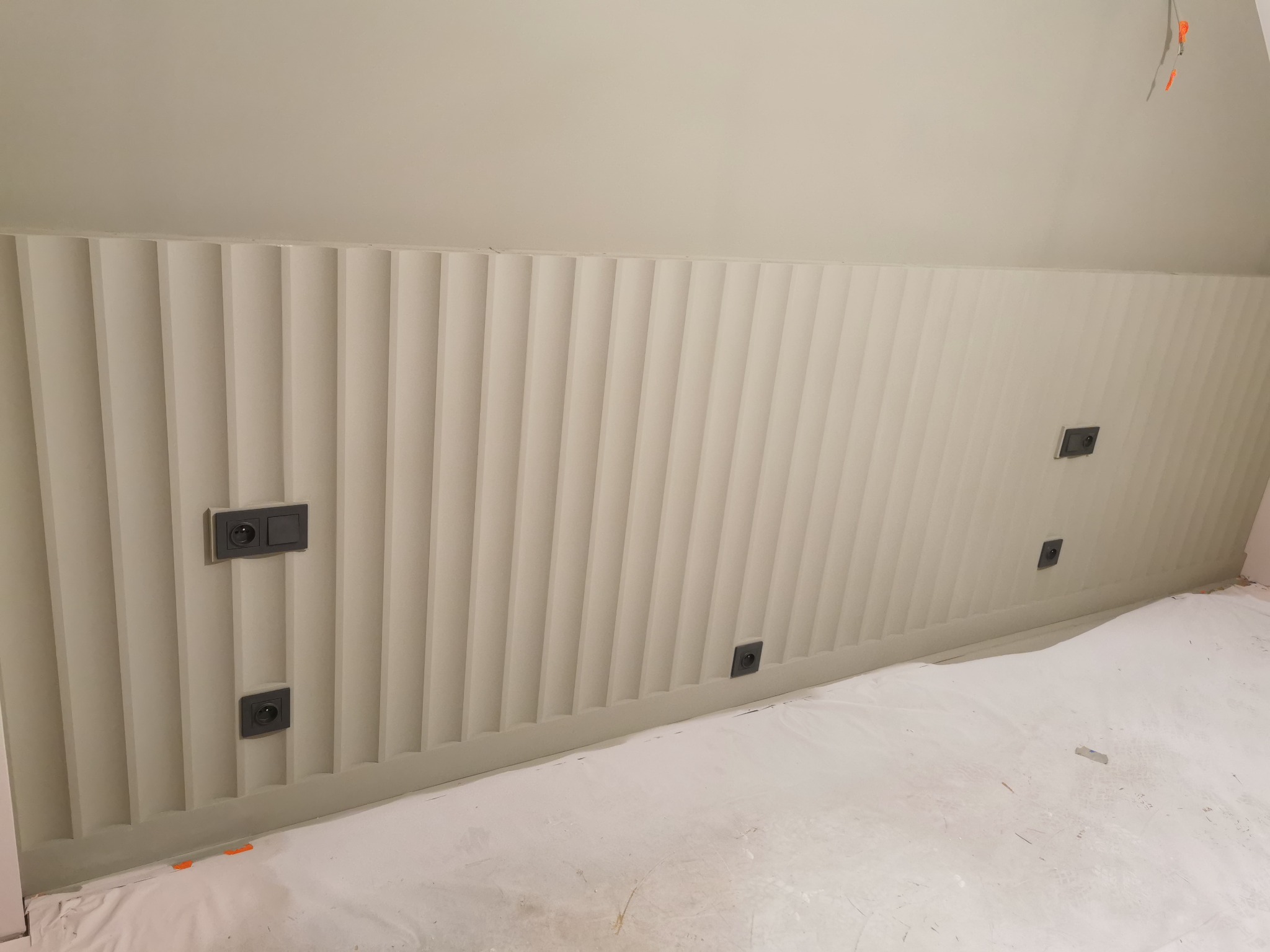 Ściana w odcieniach bieli z dekoracyjnymi pionowymi panelami i gniazdkami elektrycznymi. Podłoga zabezpieczona folią malarską.