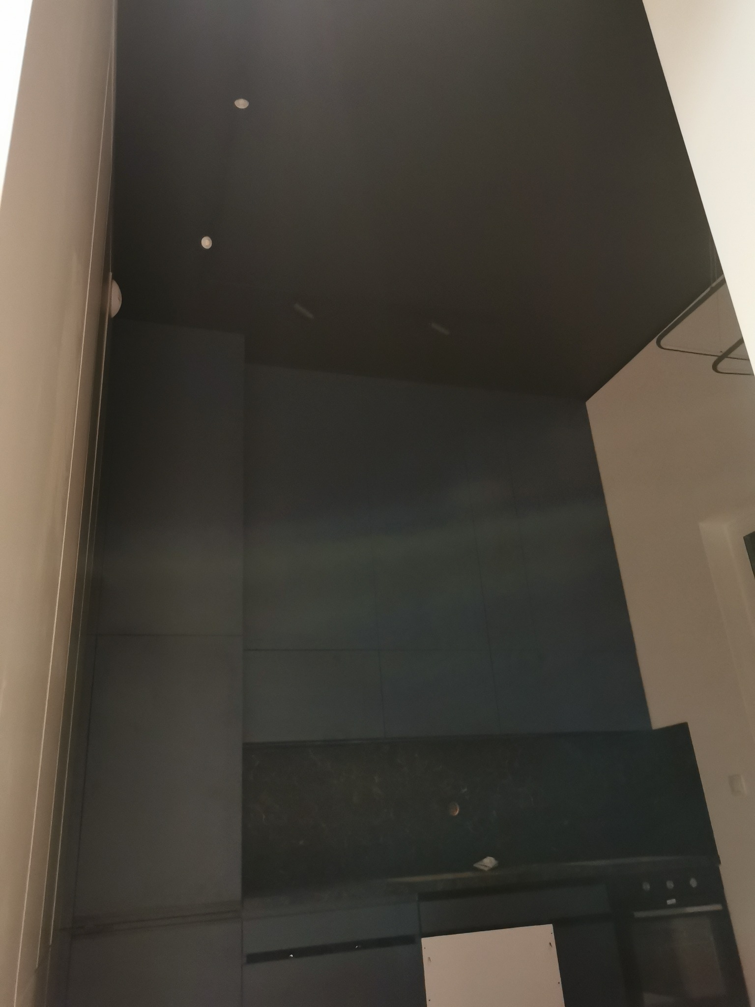 Ciemna, wykończona kuchnia z czarnym sufitem i zabudową meblową. Widoczne oświetlenie punktowe i fragment białej ściany. Ujęcie z dołu.