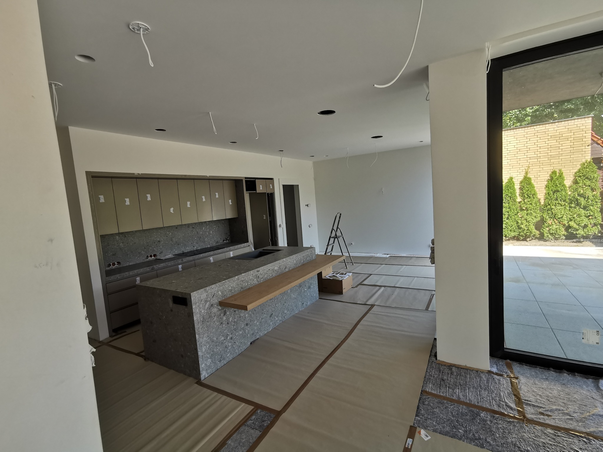 Nowoczesna kuchnia w trakcie wykończenia: wyspa z szarego kamienia, jasne ściany, zabezpieczona podłoga, surowe kable oświetleniowe, drabina w tle, widok na zieleń za oknem.