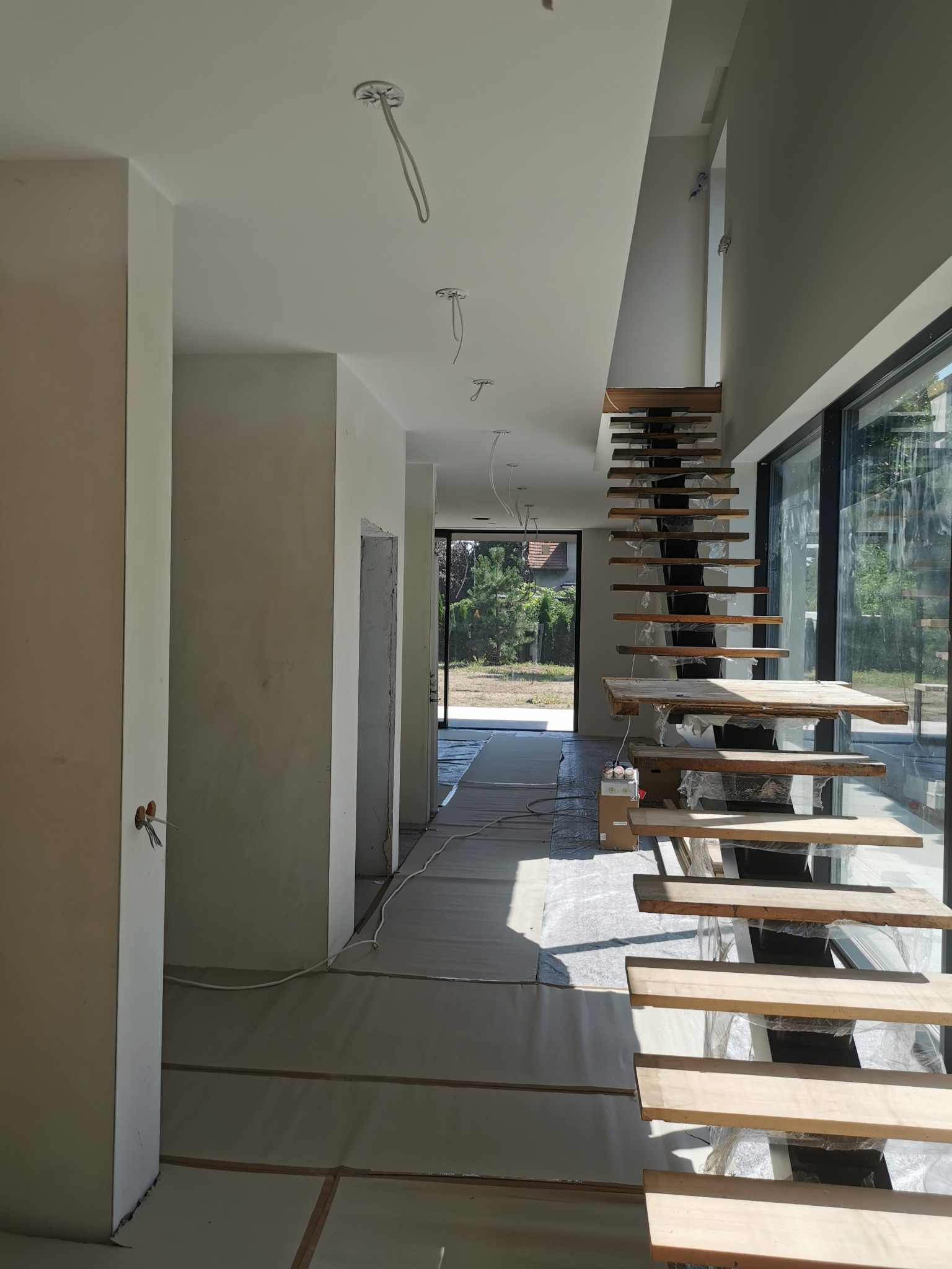Wnętrze w trakcie wykańczania: surowe drewniane schody wiszące, zabezpieczona podłoga, świeżo pomalowane ściany i przewody elektryczne zwisające z sufitu.