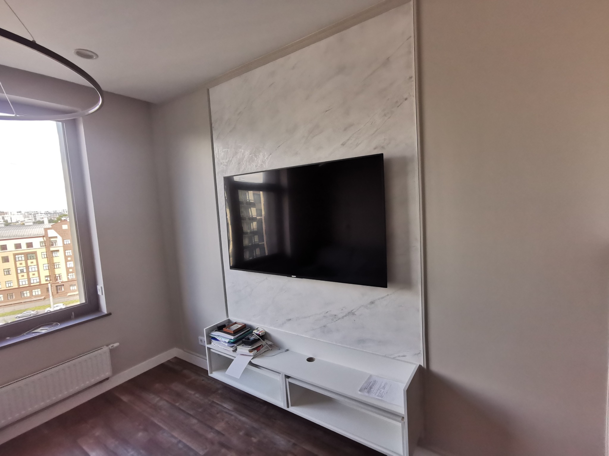 Wykończona ściana z efektem marmuru, na której zawieszony jest telewizor. Pod telewizorem biała szafka RTV. Widok na fragment pokoju z oknem i grzejnikiem.