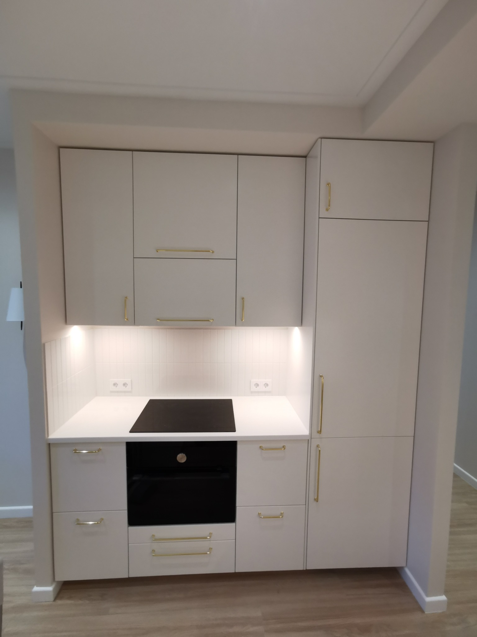 Kompaktowa, jasna zabudowa kuchenna z białymi frontami i złotymi uchwytami. Minimalistyczny design, oświetlenie LED podszafkowe, widoczne gniazdka elektryczne.