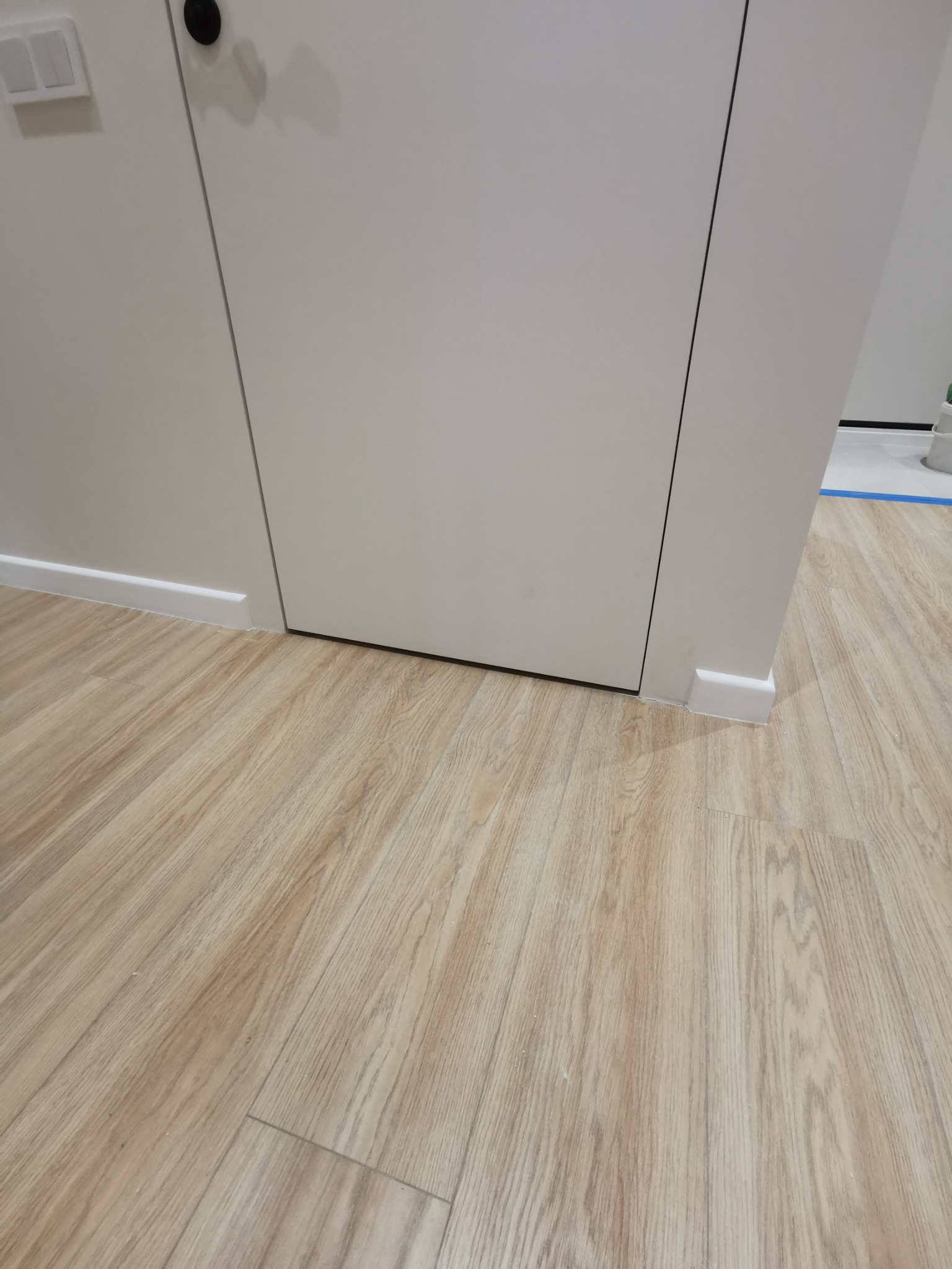 Jasne panele podłogowe ułożone wzdłuż białej ściany z drzwiami. Widoczne listwy przypodłogowe. Minimalistyczny design wnętrza.