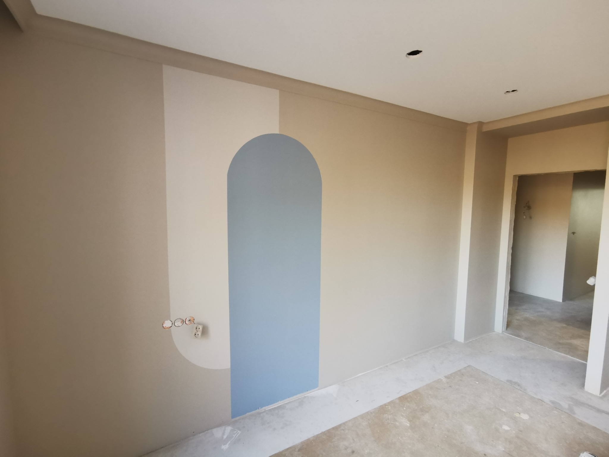 Wnętrze po odświeżeniu we Wrocławiu. Ściana z geometrycznym, niebieskim wzorem łuku, jasne kolory, widoczne gniazdka elektryczne i surowa podłoga.