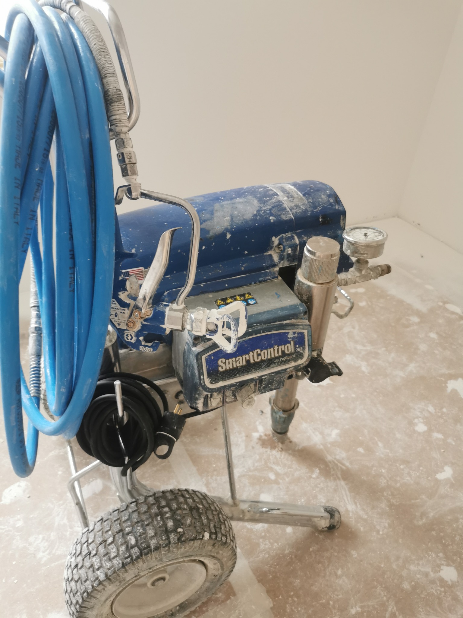 Profesjonalny agregat malarski SmartControl z niebieskim wężem, używany podczas prac wykończeniowych, z widocznymi śladami farby na podłodze i urządzeniu.
