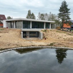 VANBLOK SPÓŁKA Z OGRANICZONĄ ODPOWIEDZIALNOŚCIĄ - Dom jednorodzinny w stanie surowym zamkniętym z dużymi oknami, usytuowany na wzgórzu z widokiem na staw, Gdańsk. Widoczne elementy betonowe i piaskowe podłoże.