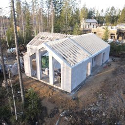 VANBLOK SPÓŁKA Z OGRANICZONĄ ODPOWIEDZIALNOŚCIĄ - Wysoka perspektywa na budowę domu z keramzytu w stanie deweloperskim, z widoczną konstrukcją dachu i ścianami z bloczków, otoczonego drzewami i innymi budynkami w tle.