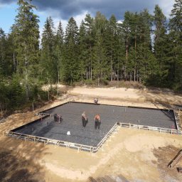 VANBLOK SPÓŁKA Z OGRANICZONĄ ODPOWIEDZIALNOŚCIĄ - Zbrojenie fundamentów pod dom jednorodzinny na leśnej działce. Widoczne szalunki, stalowe pręty i pracownicy w kombinezonach. Ujęcie z góry, w tle las iglasty.