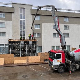 VANBLOK SPÓŁKA Z OGRANICZONĄ ODPOWIEDZIALNOŚCIĄ - Betonowanie stropu budynku komercyjnego z użyciem pompy do betonu. Widoczni pracownicy w kaskach i kamizelkach. W tle fragment budynku z oknami.