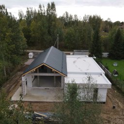 VANBLOK SPÓŁKA Z OGRANICZONĄ ODPOWIEDZIALNOŚCIĄ - Dom jednorodzinny w stanie surowym otwartym z dachem z blachy trapezowej i płaskim dachem, otoczony zielenią. Widok z góry ukazuje postęp prac budowlanych.