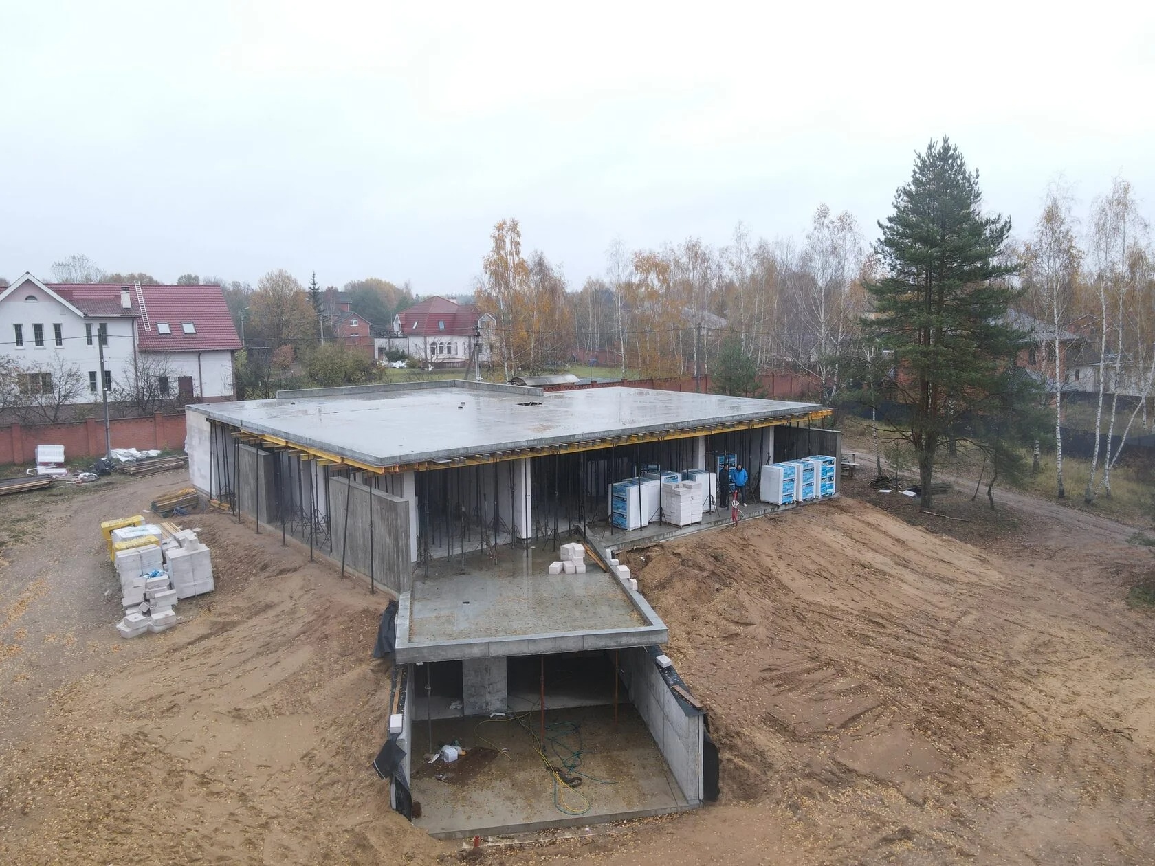 Stan surowy zamknięty domu jednorodzinnego z płaskim dachem, widok z góry. Na pierwszym planie piwnica, w tle zabudowa jednorodzinna i drzewa.