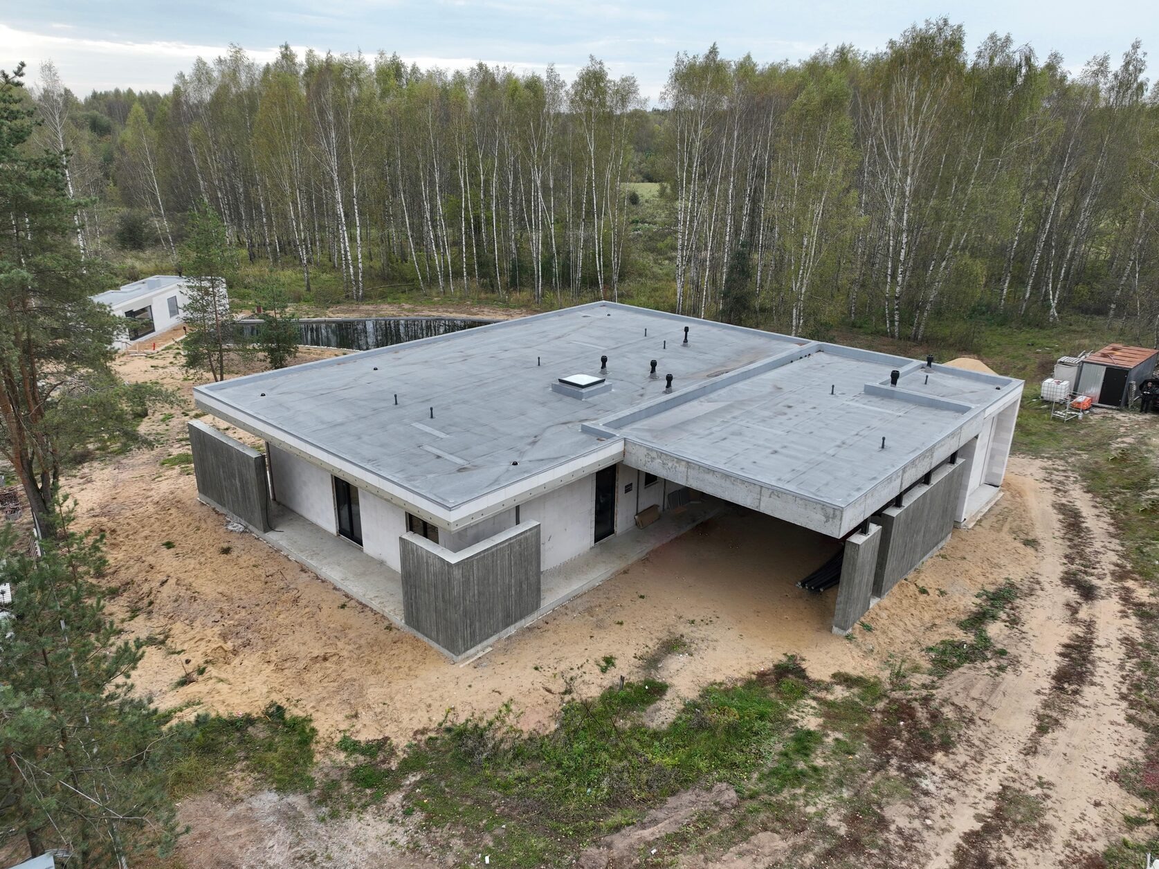 Nowoczesny dom jednorodzinny z płaskim dachem, betonowymi elementami i dużymi oknami, otoczony piaszczystym terenem i lasem. Widok z lotu ptaka.