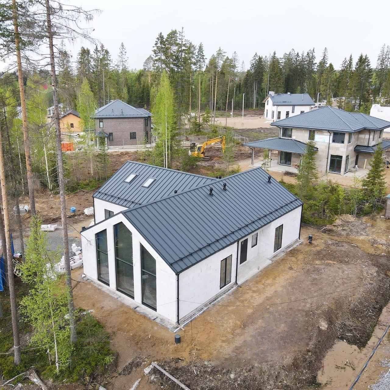 Nowoczesny dom jednorodzinny z ciemnym dachem na działce z drzewami, widziany z góry. Duże okna z czarnymi ramami. W tle inne domy i koparka.