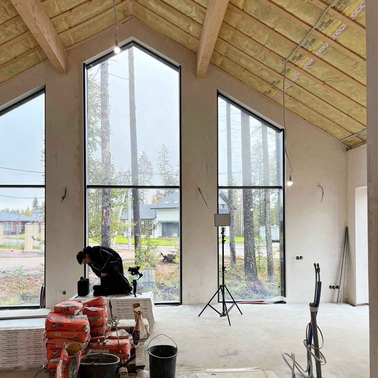 Wnętrze domu w stanie deweloperskim z dużymi oknami i widoczną izolacją dachu. Na podłodze worki z zaprawą, osoba filmująca wnętrze.