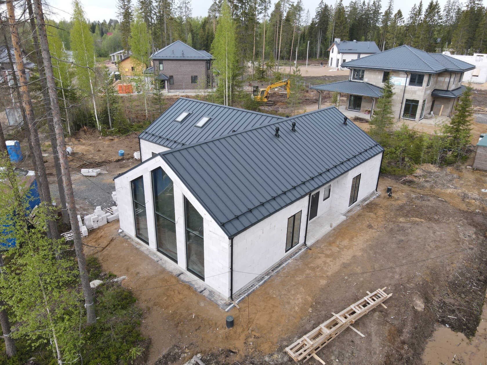 Nowoczesny dom jednorodzinny w stanie deweloperskim z ciemnym dachem na tle lasu. Widok z góry ukazuje bryłę budynku i otoczenie w naturalnym krajobrazie.