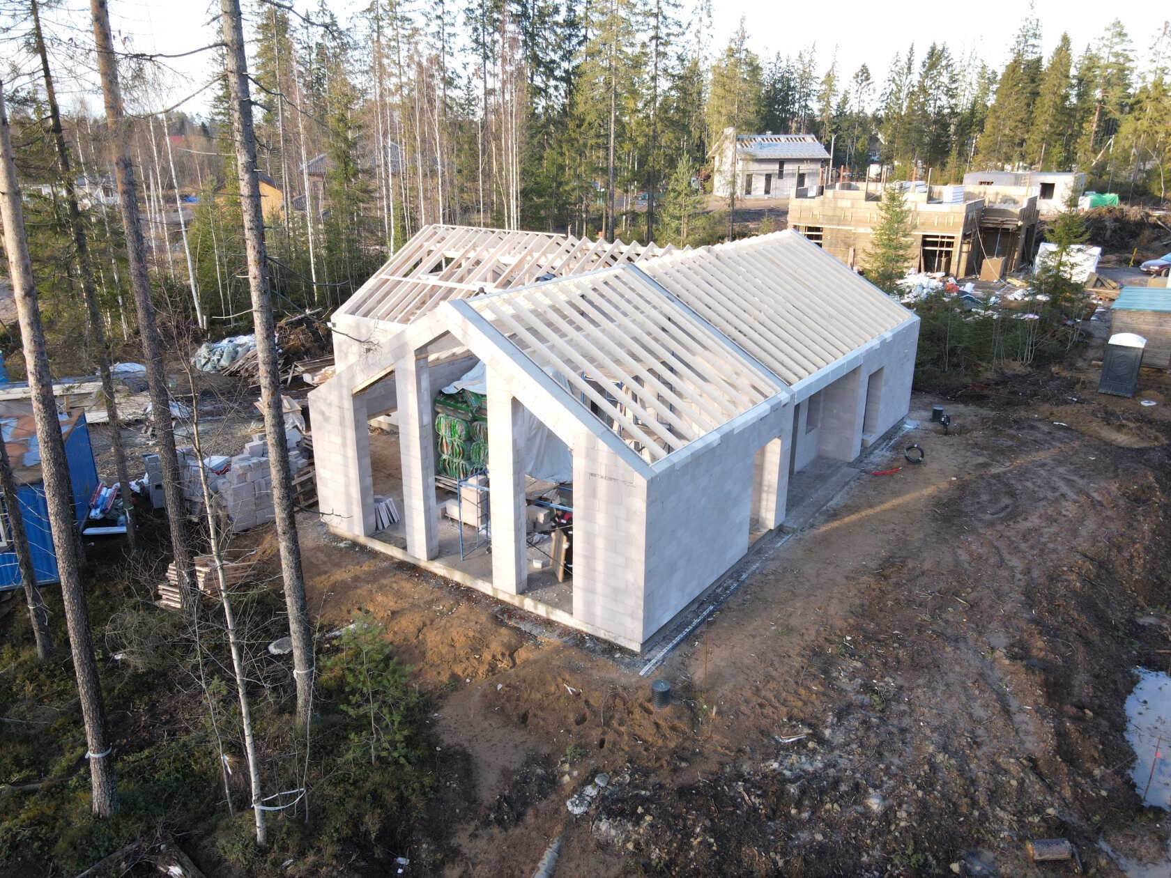 Wysoka perspektywa na budowę domu z keramzytu w stanie deweloperskim, z widoczną konstrukcją dachu i ścianami z bloczków, otoczonego drzewami i innymi budynkami w tle.