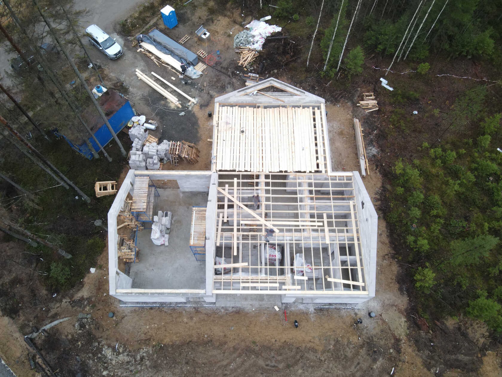 Widok z lotu ptaka na budowę domu jednorodzinnego w stanie surowym otwartym, z widocznymi fundamentami, ścianami z bloczków i konstrukcją dachu z drewnianych belek.