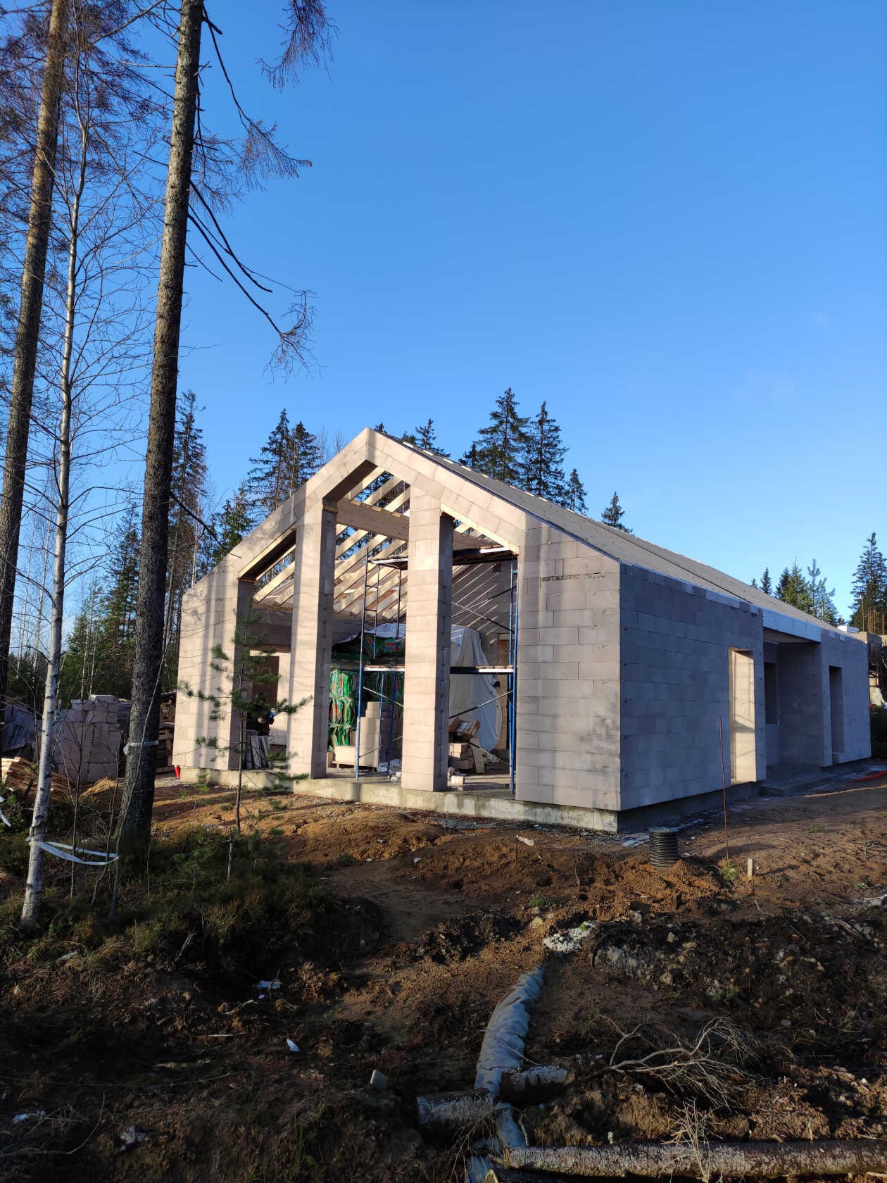 Surowy stan domu z betonu komórkowego w lesie, z widoczną konstrukcją dachu i otworami okiennymi. Wokół plac budowy z ziemią i drzewami. Słoneczny dzień.