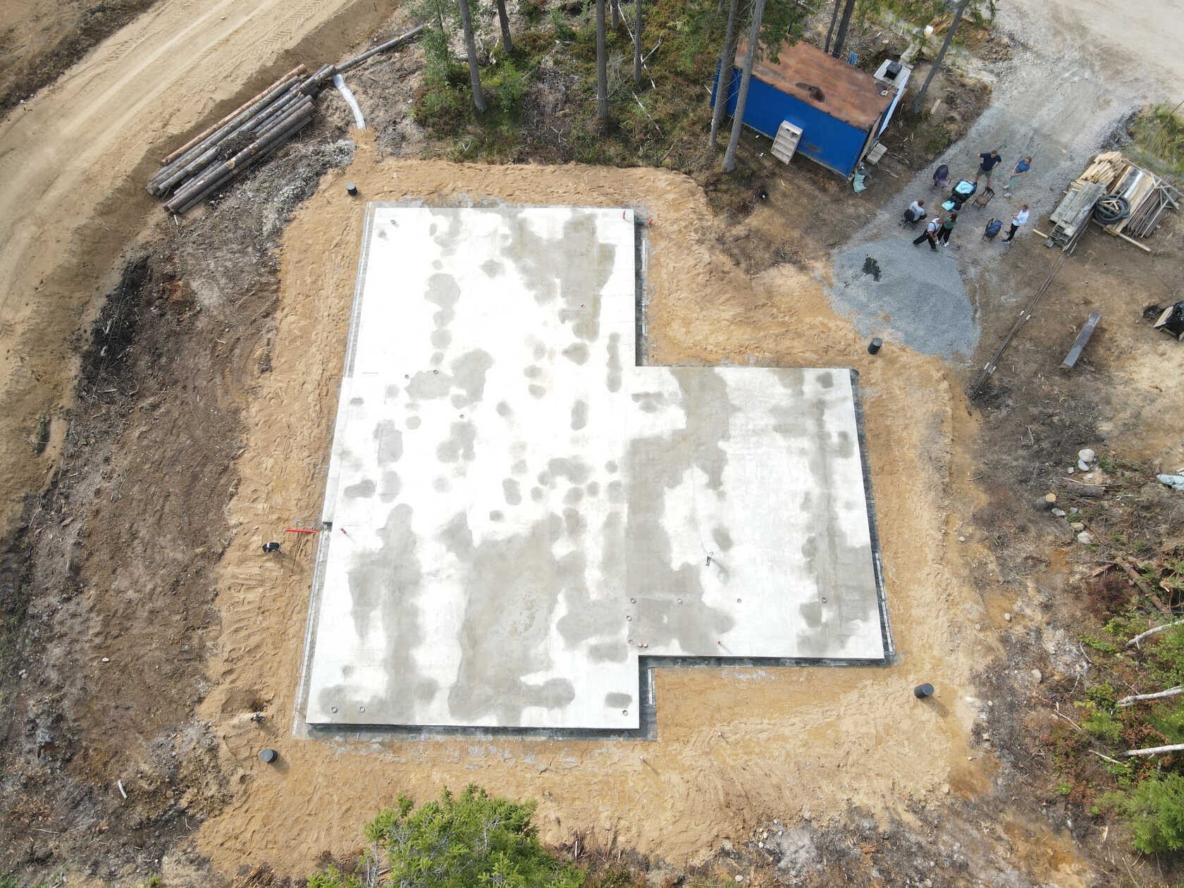 Z lotu ptaka: wylany fundament domu jednorodzinnego na piaszczystym terenie, otoczony lasem. Widoczny barak budowlany i zgromadzone drewno.