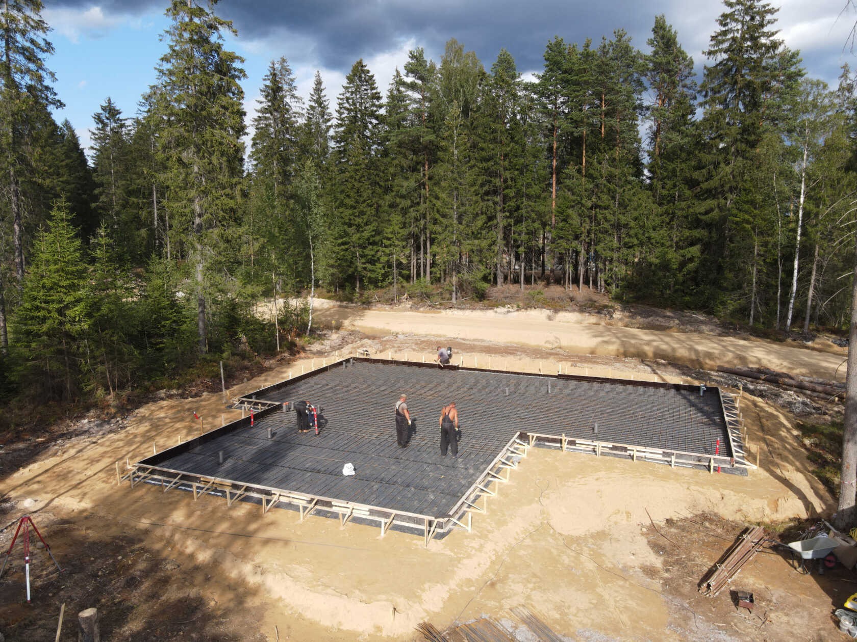 Zbrojenie fundamentów pod dom jednorodzinny na leśnej działce. Widoczne szalunki, stalowe pręty i pracownicy w kombinezonach. Ujęcie z góry, w tle las iglasty.