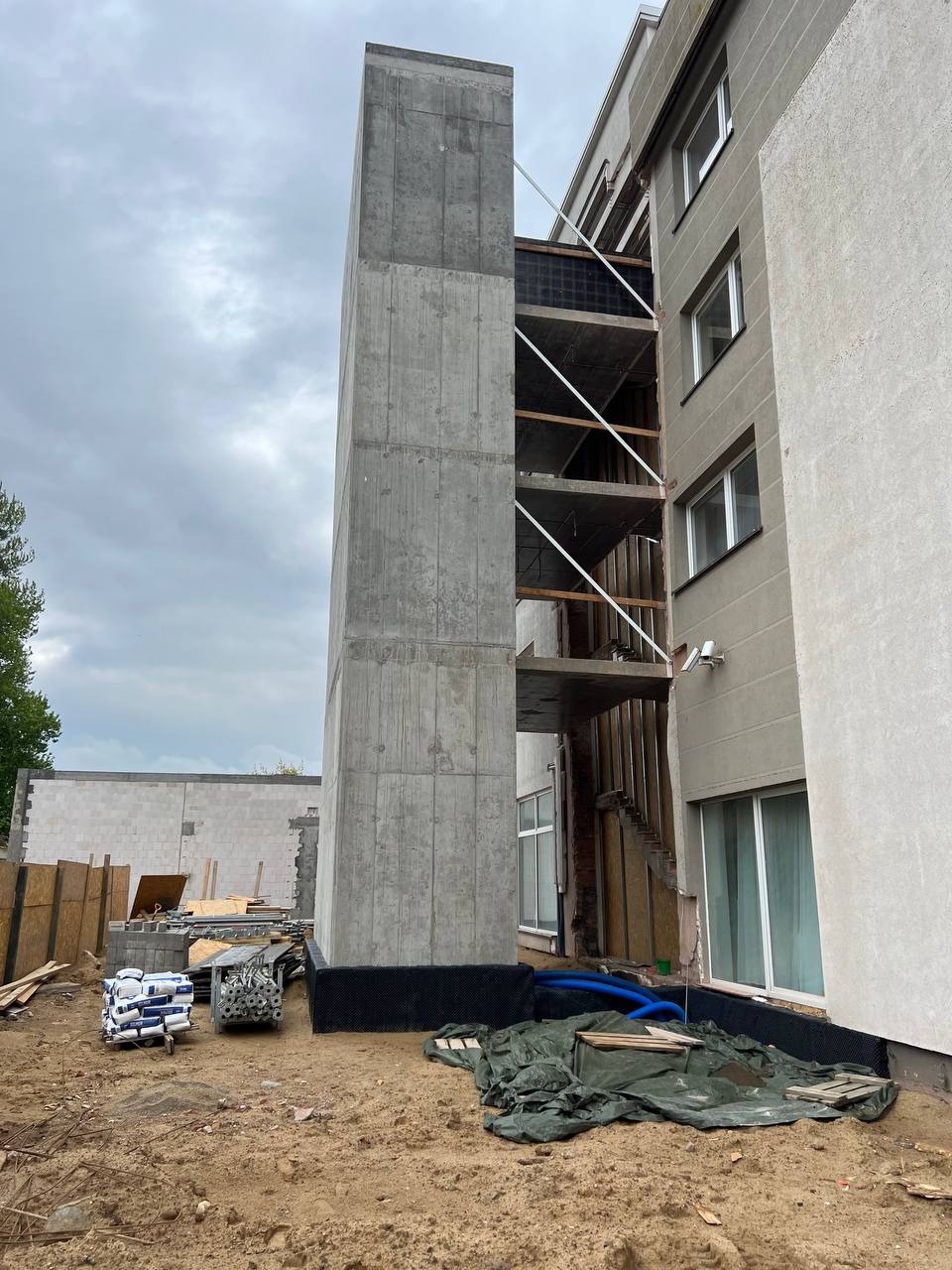 Betonowy szyb windowy w trakcie budowy, przylegający do istniejącego budynku. Widoczne materiały budowlane i elementy konstrukcyjne. Ujęcie z dołu, pochmurne niebo.