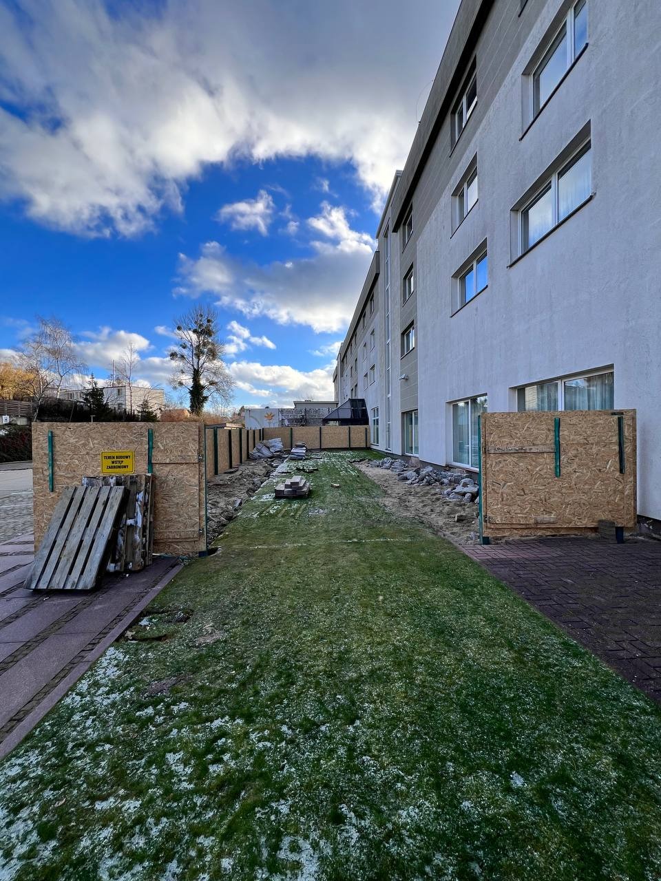 Budowa hotelu w toku: widok na teren budowy z zabezpieczeniami, częściowo wykopany teren, przygotowania do fundamentów, budynek hotelowy w tle, pochmurne niebo.