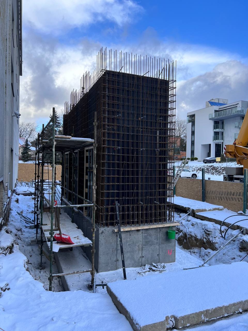 Zimowy kadr z budowy hotelu: zbrojenie ścian na fundamencie, rusztowania, śnieg. Widoczne elementy konstrukcyjne i otoczenie placu budowy.