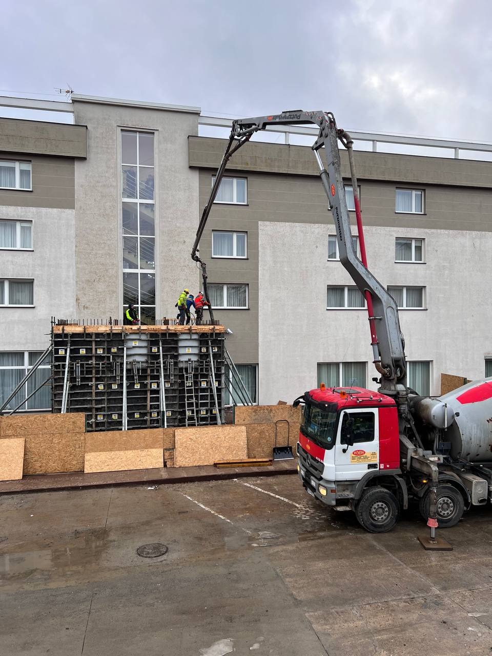 Betonowanie stropu budynku komercyjnego z użyciem pompy do betonu. Widoczni pracownicy w kaskach i kamizelkach. W tle fragment budynku z oknami.