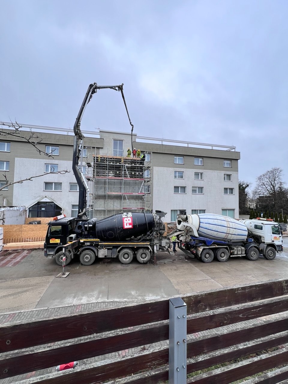 Betonowanie stropu hotelu z użyciem pompy do betonu. Na rusztowaniu pracownicy w kaskach. W tle budynek hotelu z oknami. Dwa betonomieszarki na placu budowy.