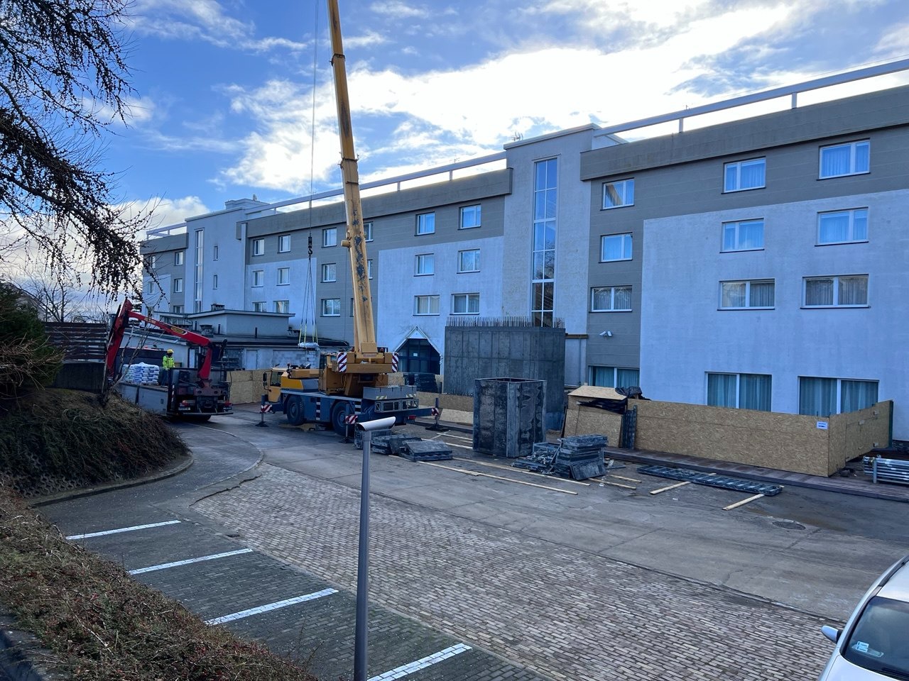 Budowa hotelu: żuraw podnosi element konstrukcyjny obok betonowego szalunku. Widoczny fragment elewacji budynku i materiały budowlane na placu.