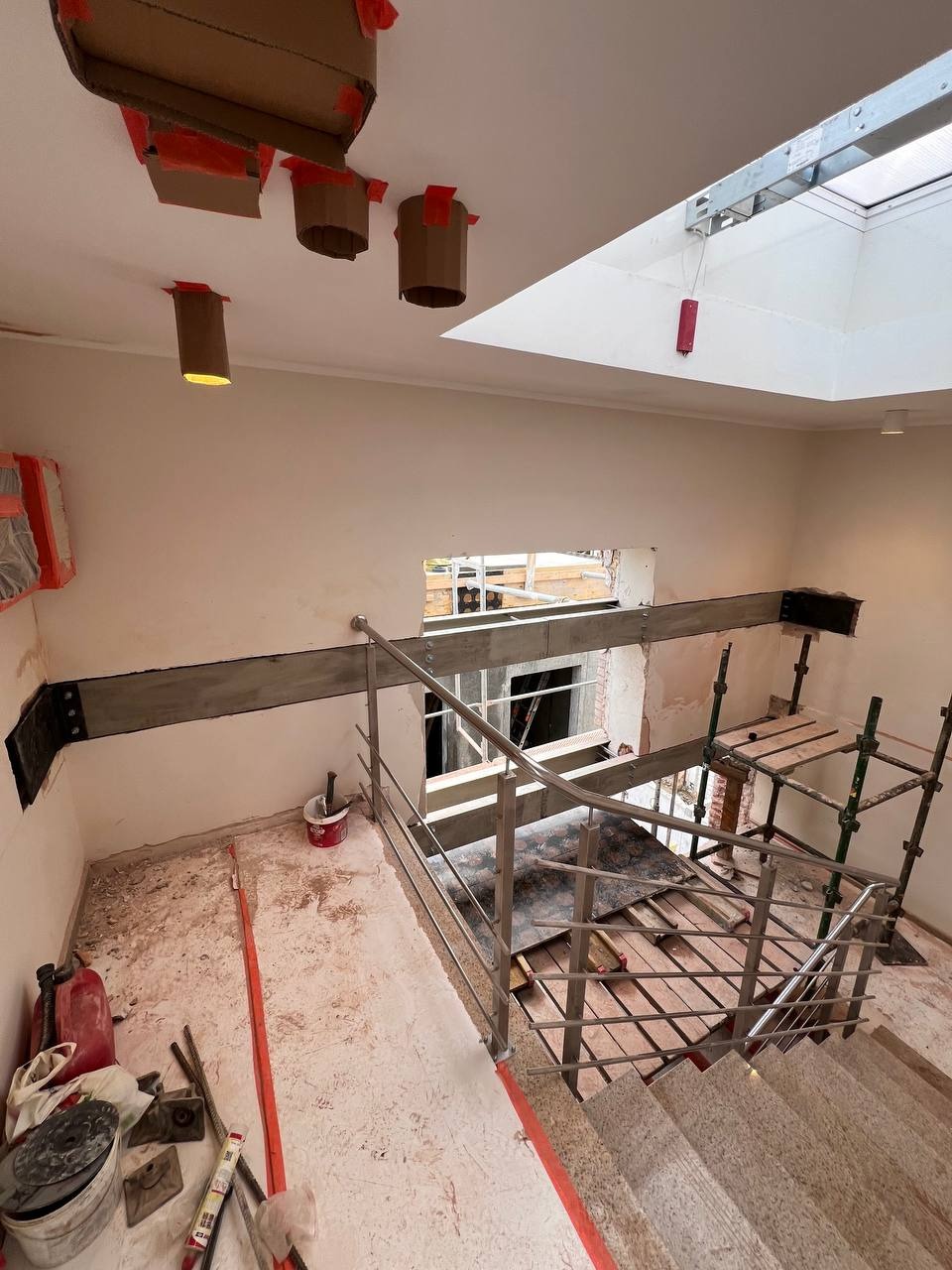 Wnętrze budynku w trakcie wzmacniania konstrukcji, widoczne stalowe belki, prowizoryczne zabezpieczenia oświetlenia i schody w budowie. Prace remontowe w obiekcie komercyjnym.