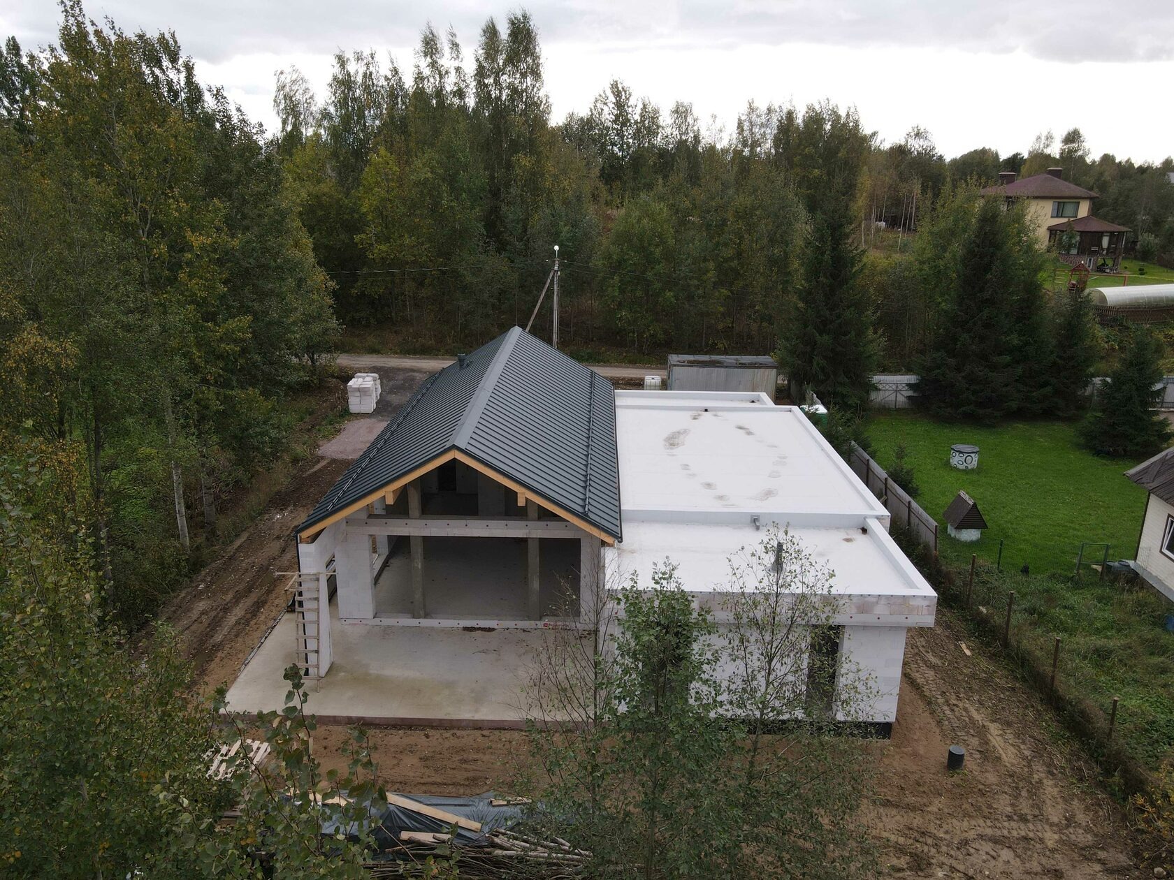 Dom jednorodzinny w stanie surowym otwartym z dachem z blachy trapezowej i płaskim dachem, otoczony zielenią. Widok z góry ukazuje postęp prac budowlanych.