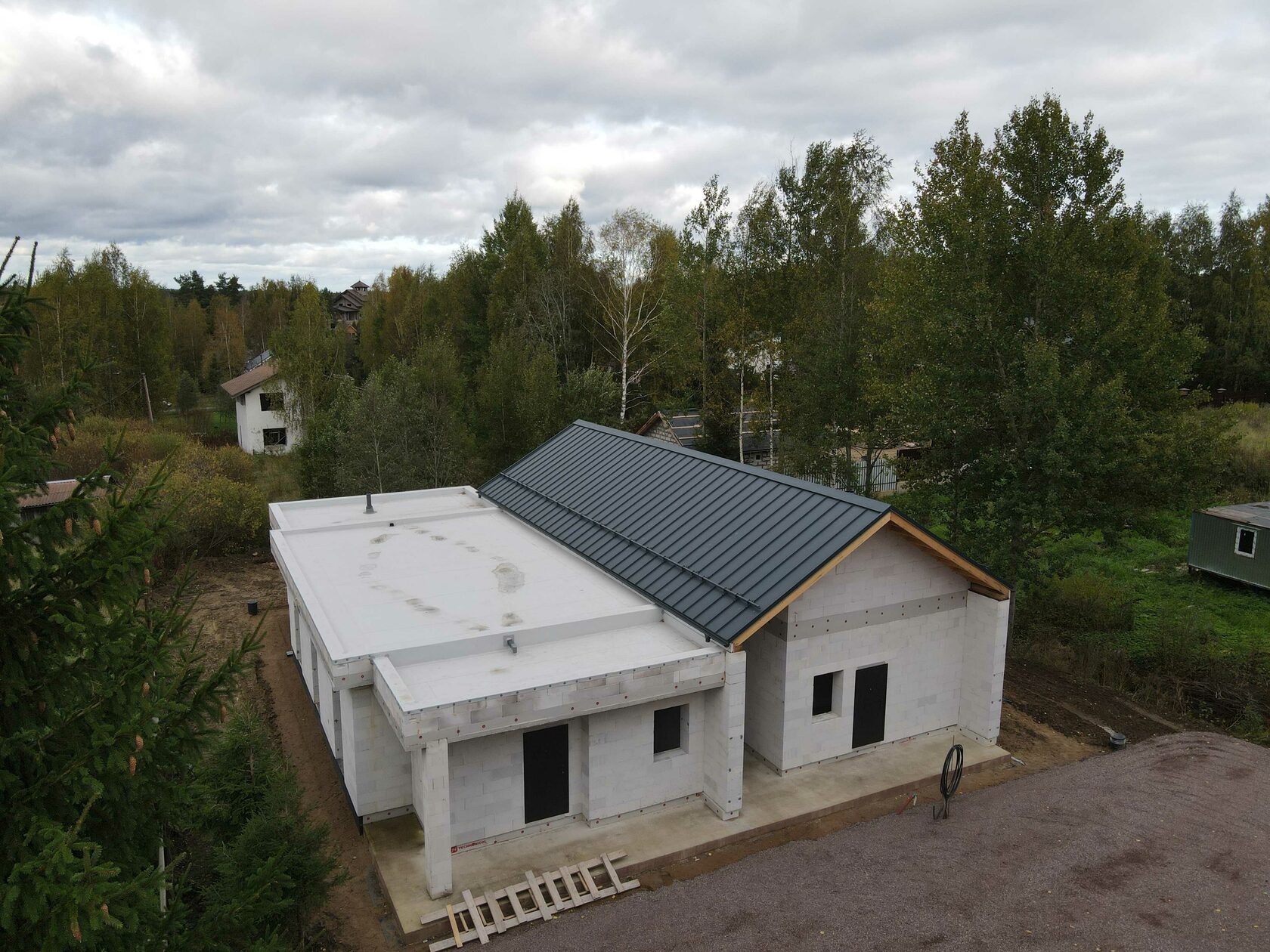 Dom jednorodzinny w stanie surowym otwartym z dachem z blachy i płaskim dachem, otoczony drzewami, widziany z góry. Na ziemi leżą drewniane schody.