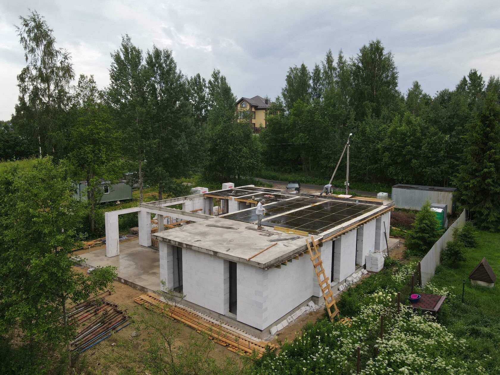 Dom jednorodzinny w stanie surowym otwartym, z białych bloczków, z widocznym zbrojeniem stropu. W tle zieleń drzew i zabudowa jednorodzinna. Ujęcie z góry.