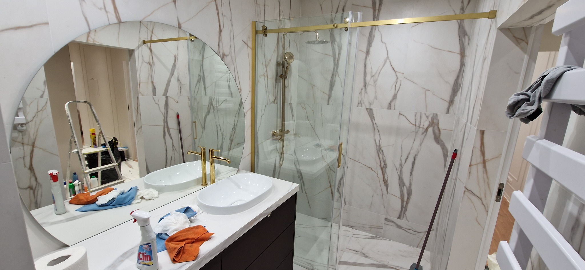 Nowoczesna łazienka z marmurowymi płytkami, złotą armaturą i kabiną prysznicową typu walk-in, widoczne środki czystości i drabina malarska sugerują niedawne zakończenie prac.