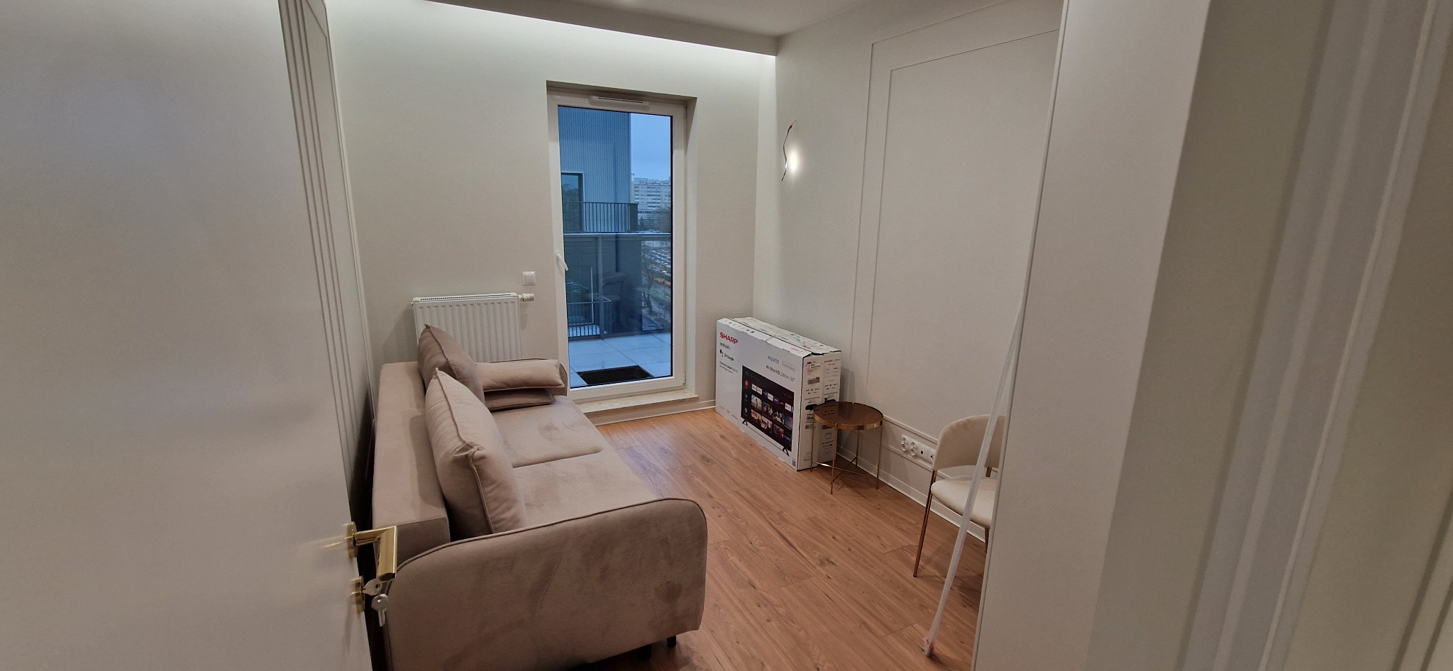 Jasne wnętrze pokoju z beżową sofą, grzejnikiem pod oknem balkonowym, kartonem po telewizorze i krzesłem. Na podłodze panele w odcieniach brązu. Widok zza uchylonych drzwi.