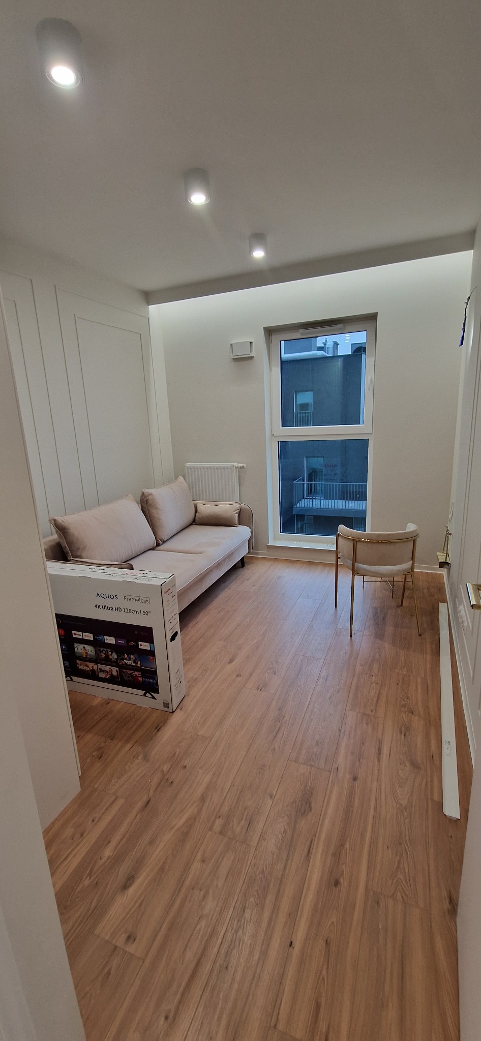 Jasne wnętrze pokoju z drewnianą podłogą, beżową sofą, krzesłem na złotych nogach i zapakowanym telewizorem Aquos 50 cali, widoczne okno z widokiem na balkon sąsiedniego budynku.